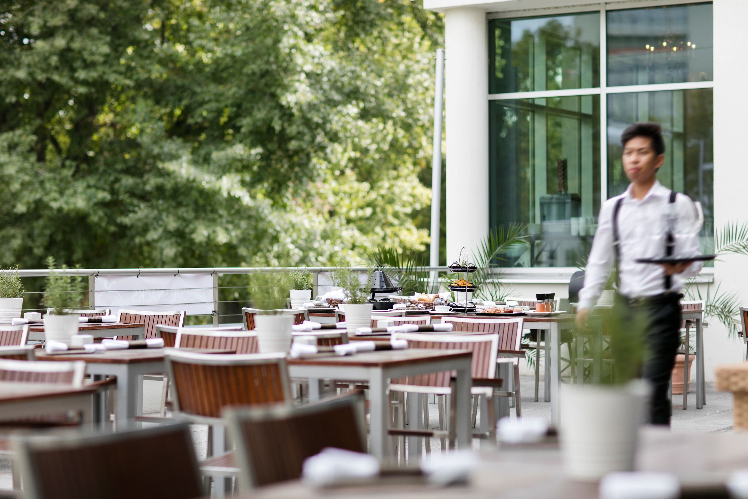 Ein Straßencafé mit Tischen und Stühlen, ein Kellner geht vorbei, und im Hintergrund ein Gebäude mit großen Fenstern.
