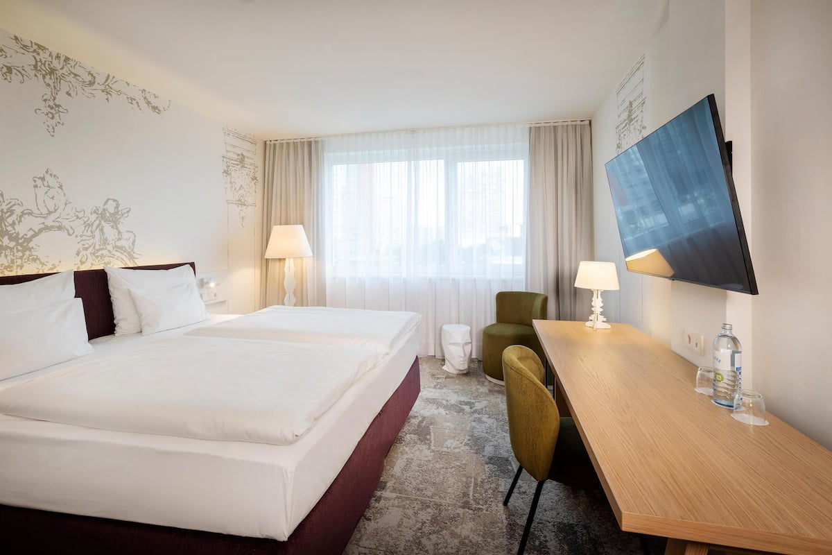 Habitación de hotel con una cama grande, dos mesitas de noche, un escritorio, una silla, un televisor y una ventana con cortinas.