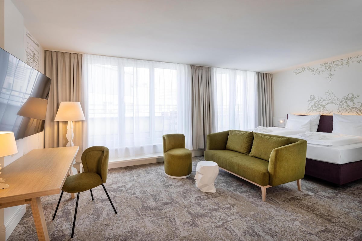 Una habitación de hotel con una cama, un sofá verde, una mesita redonda pequeña, una lámpara y un espejo grande.