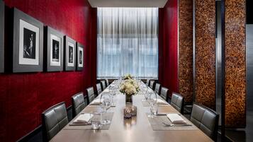 A conference room with a long table, chairs, framed pictures on the wall, and a large window with curtains.