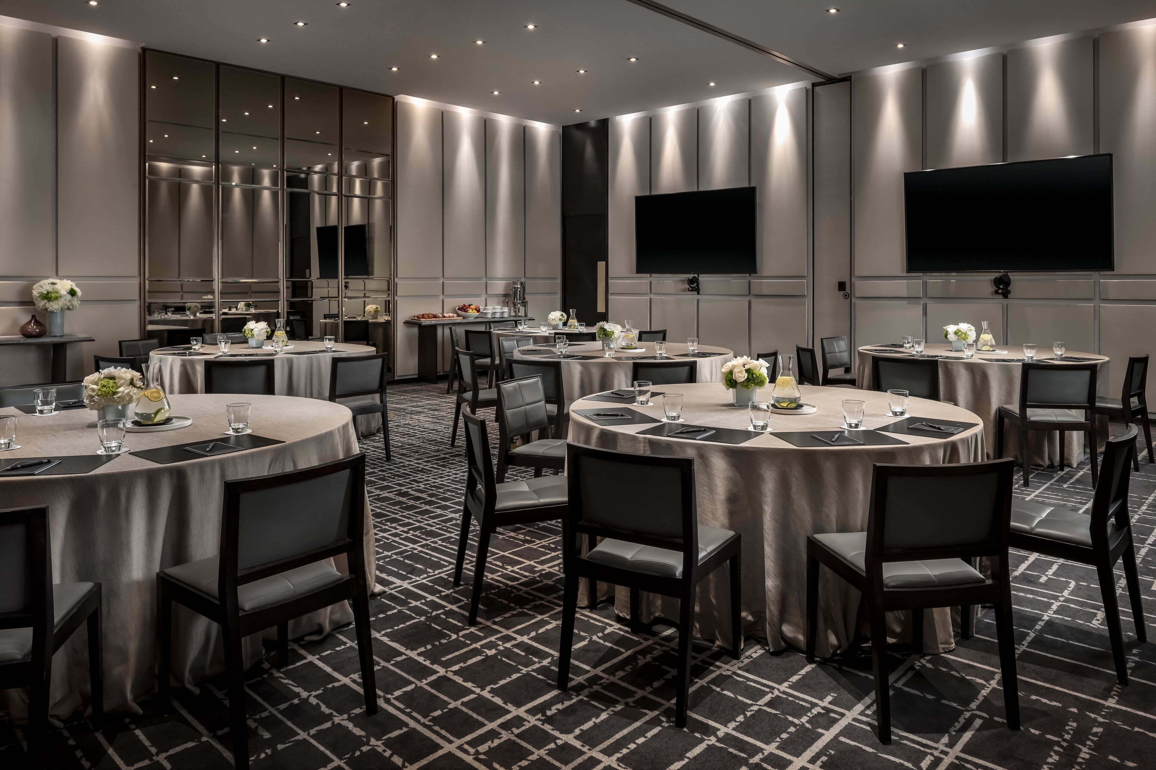 Une salle de conférence moderne, dotée de tables rondes, de chaises noires et d’un grand téléviseur à écran plat fixé au mur.