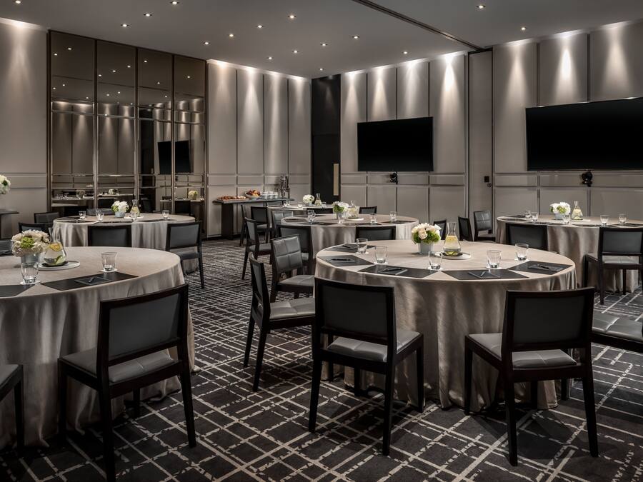 Une salle de conférence moderne, dotée de tables rondes, de chaises noires et d’un grand téléviseur à écran plat fixé au mur.