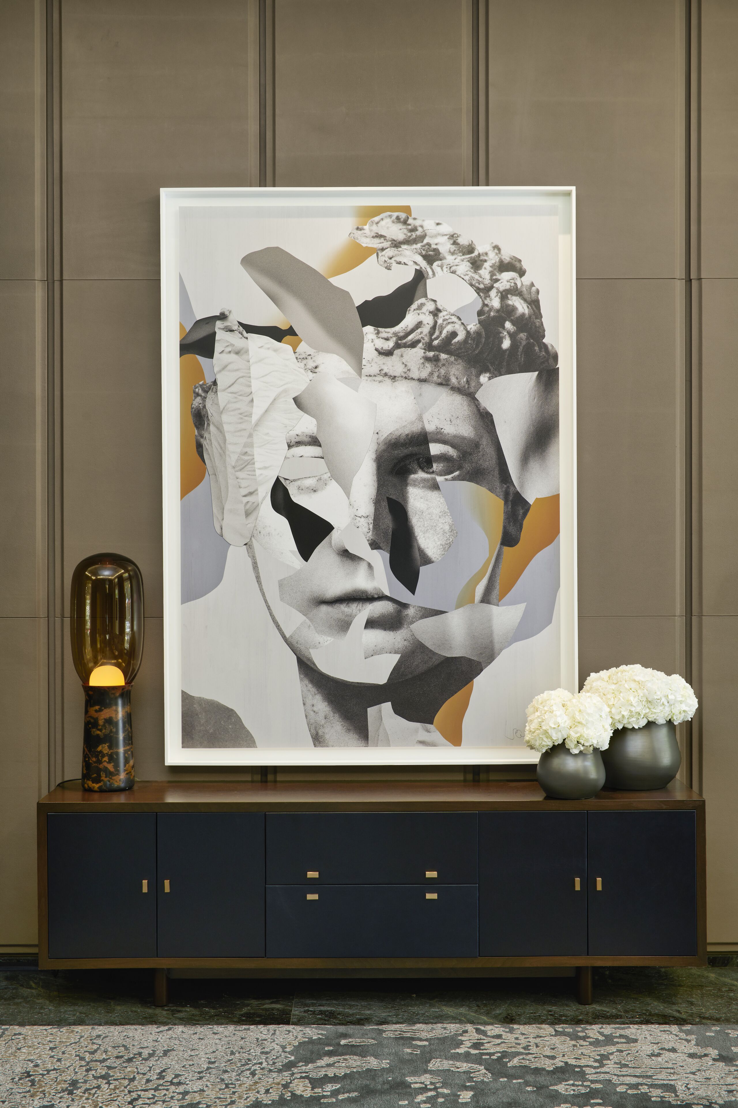 A framed abstract artwork on a dark blue cabinet with gold handles, a decorative vase with white flowers, and a lit lamp.