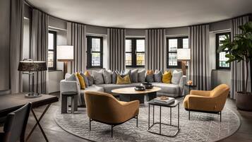 Un salon moderne avec un canapé incurvé, deux fauteuils, une table basse et une table d’appoint. De grandes fenêtres sont agrémentées de rideaux, et on y trouve une lampe ainsi qu’une plante en pot.