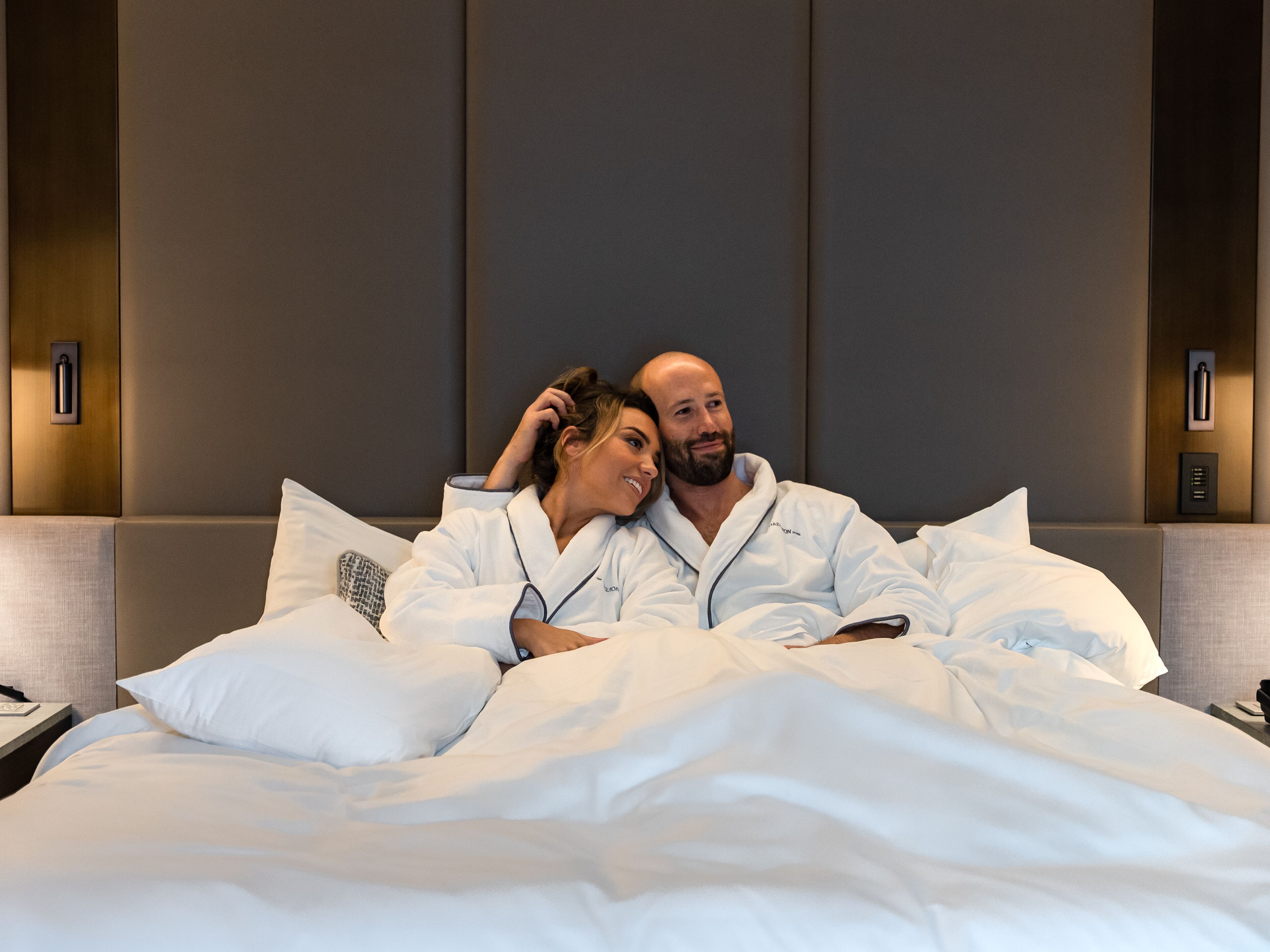 A modern hotel room with a large bed, a headboard with built-in lighting, and a bedside table with a lamp.