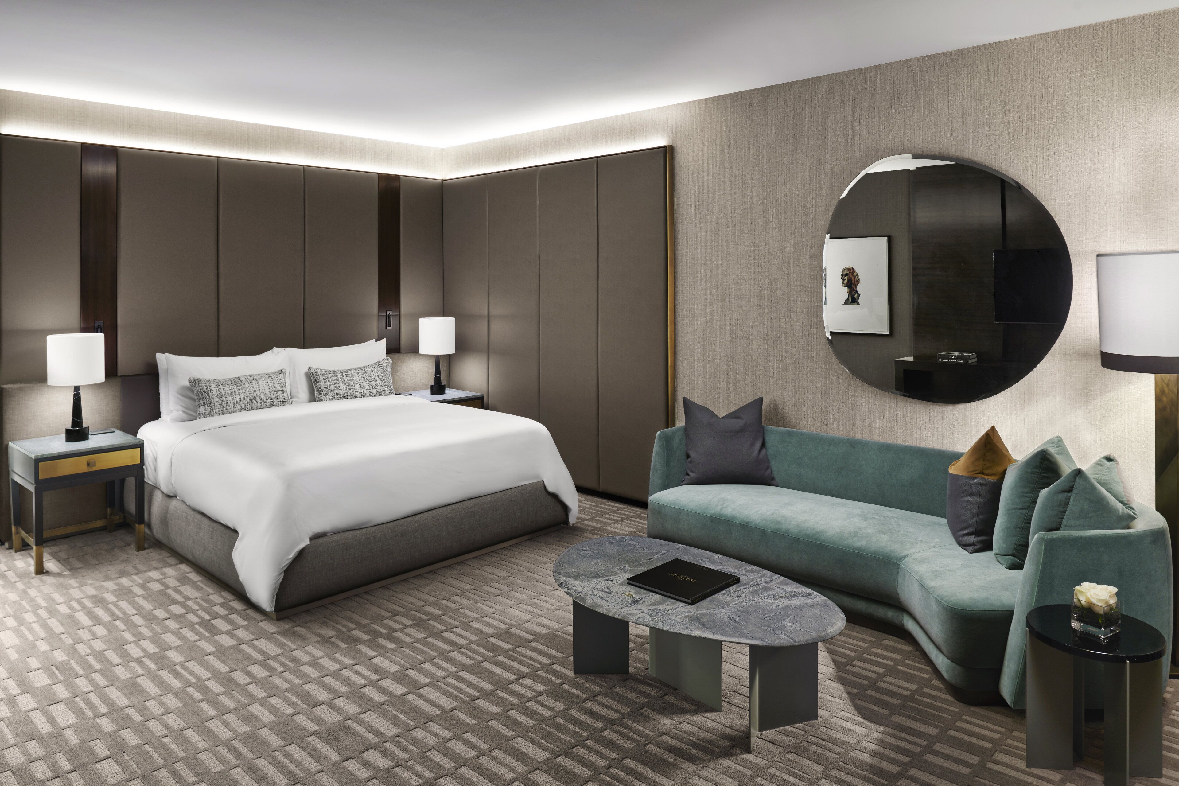 A modern hotel room with a large bed, a sofa, a coffee table, and a round mirror.