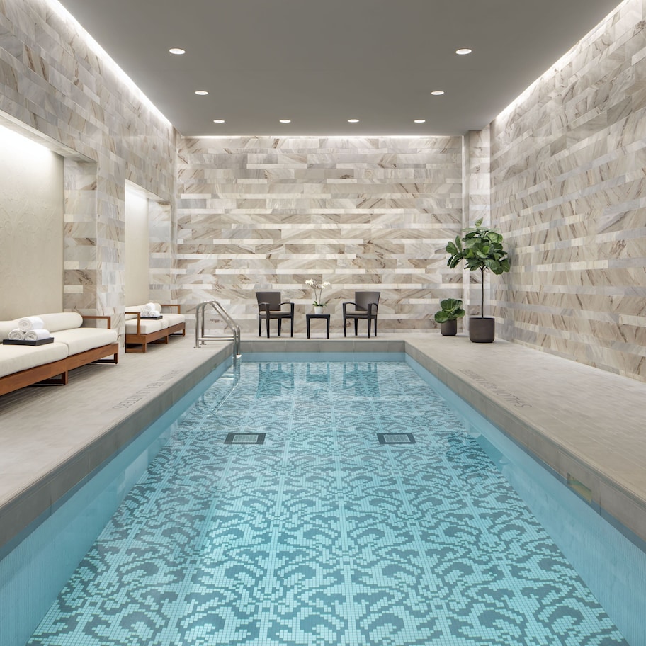 A modern indoor pool with a patterned floor, seating area with chairs and a sofa, and a wall with a herringbone pattern.