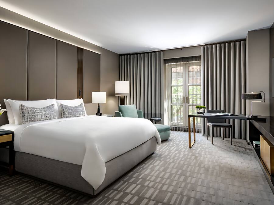 A modern hotel room with a large bed, two bedside tables with lamps, a desk with a chair, and a TV on the wall.