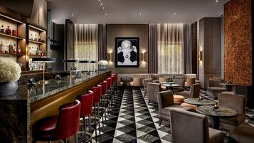 A bar area with a marble countertop, red bar stools, and a wall-mounted liquor display. There are round tables and chairs arranged for dining or socializing.