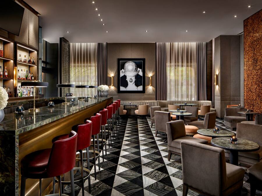 A bar area with a marble countertop, red bar stools, and a wall-mounted liquor display. There are round tables and chairs arranged for dining or socializing.