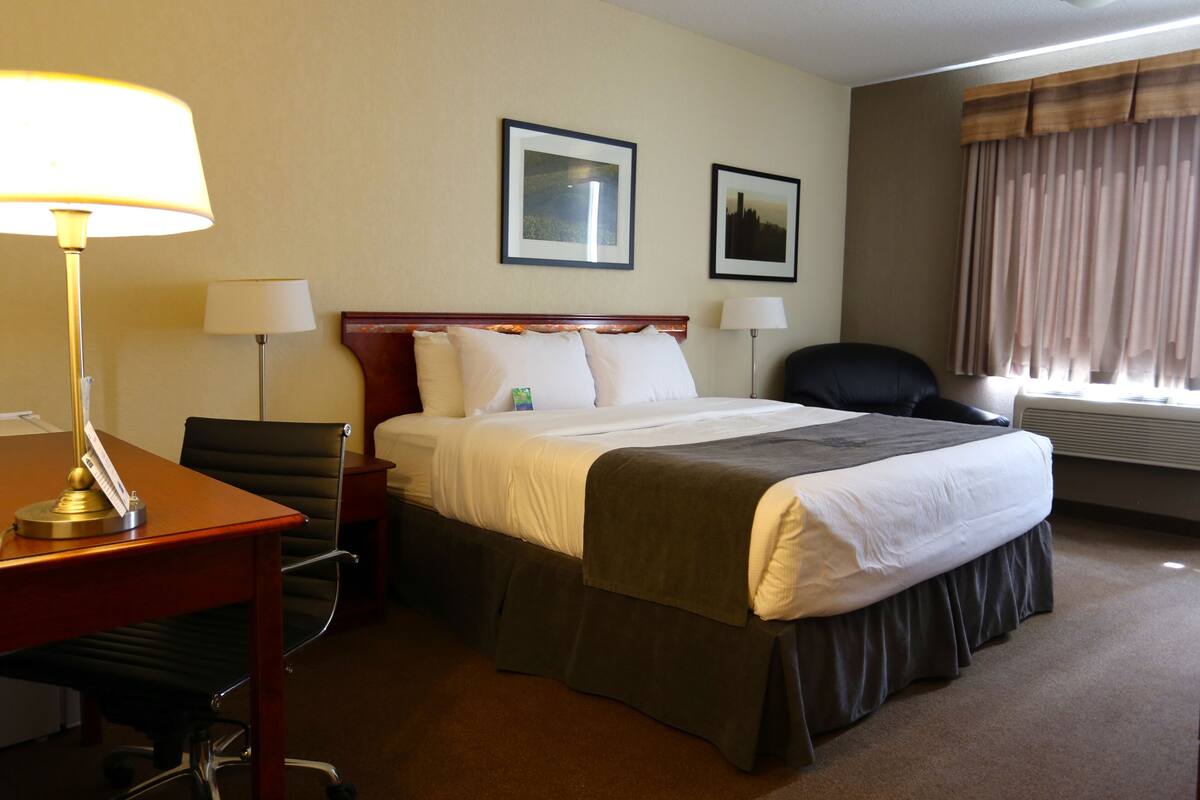 A hotel room with a bed, two bedside lamps, a desk with a lamp, a chair, and two framed pictures on the wall.