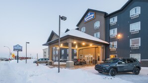 A hotel with a covered entrance, a parking lot with several cars, and a sign indicating the hotel name.