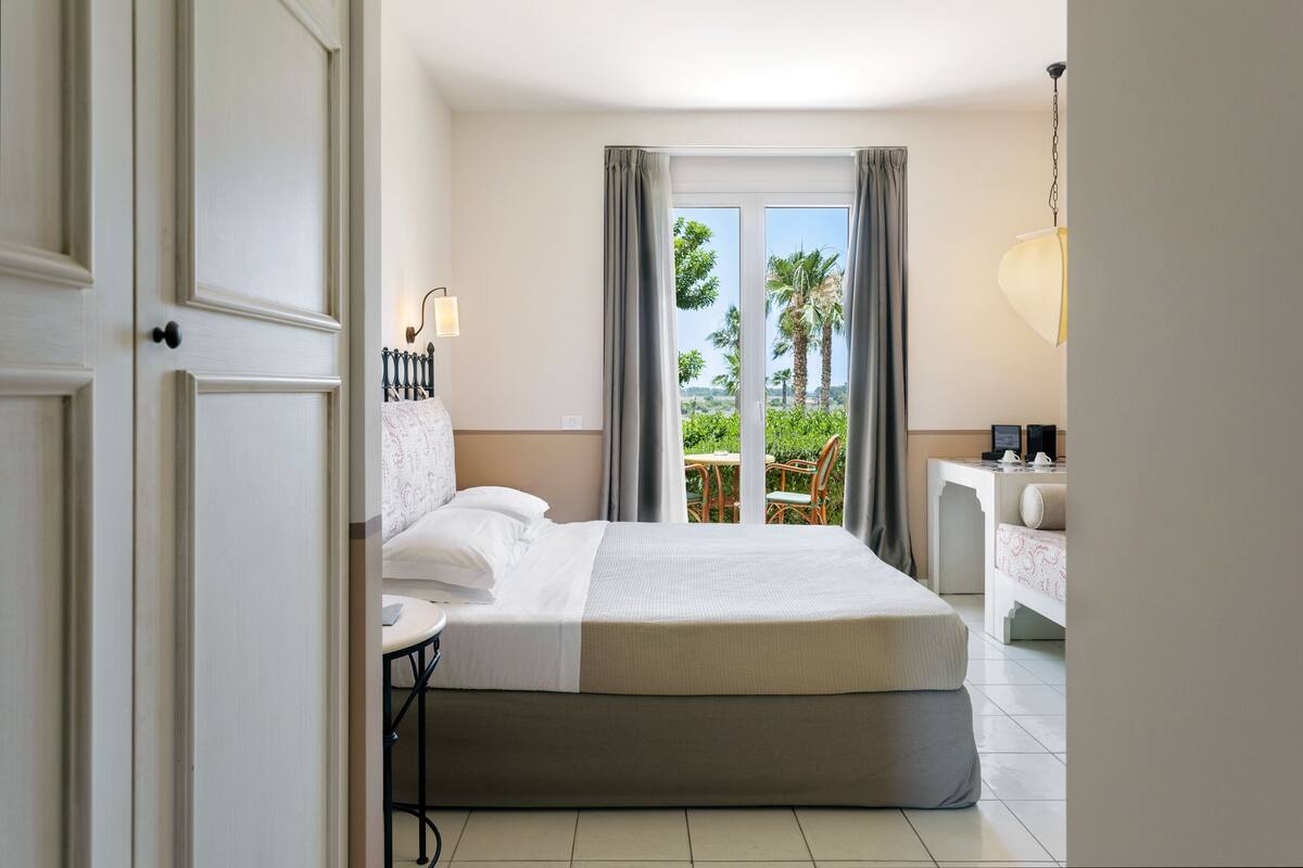 Une chambre d’hôtel avec un lit, une petite table, une chaise, un bureau et une vue sur les palmiers par la fenêtre.