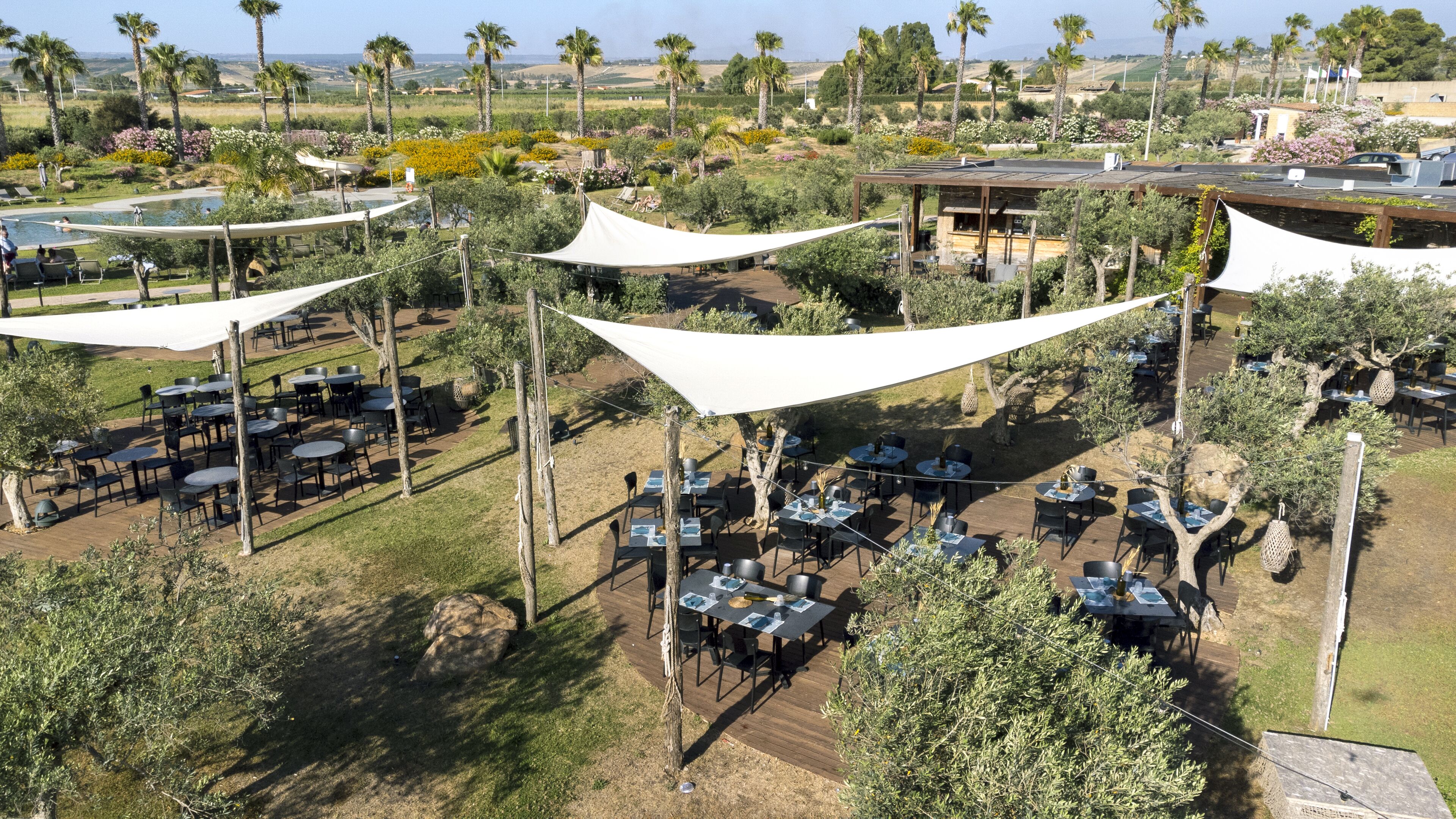 Un área de comedor al aire libre con mesas y sillas bajo grandes toldos blancos, rodeada de follaje y palmeras.