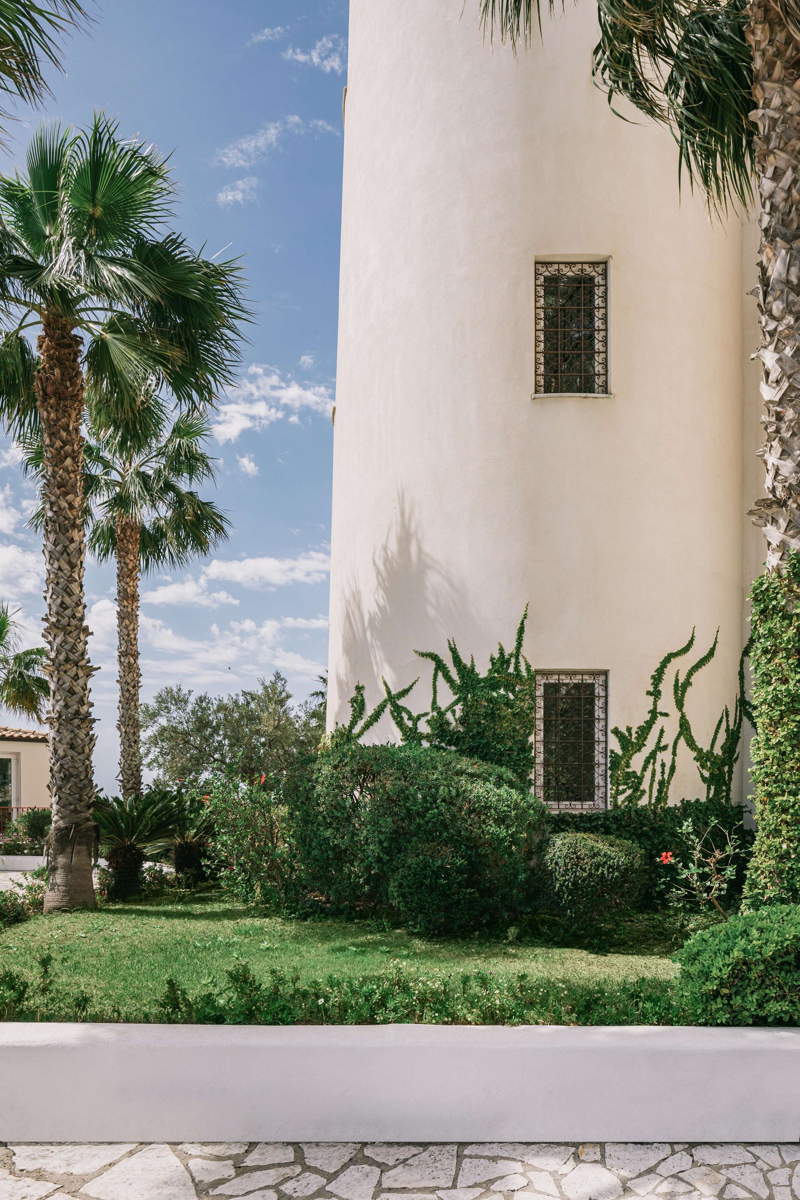 A tall, cylindrical building with two windows, surrounded by lush greenery and palm trees.