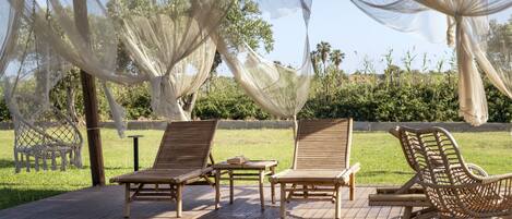Un espacio al aire libre cubierto con sillones de madera y una mesa, rodeado de follaje y un cielo despejado.