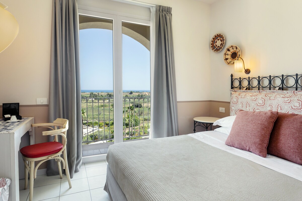 A hotel room with a bed, a desk, a chair, and a view of the countryside through large windows.