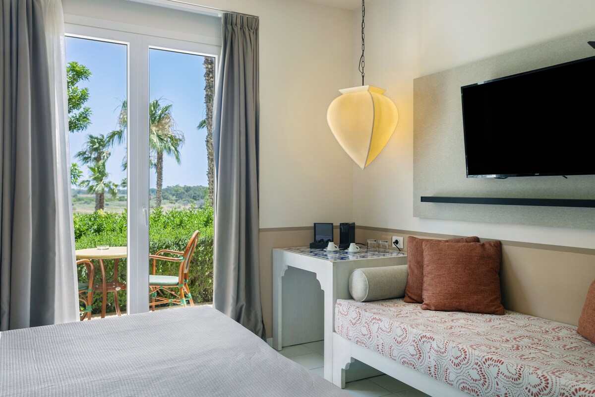 A hotel room with a bed, a small table with a coffee maker, a TV mounted on the wall, and a balcony with a table and chairs.