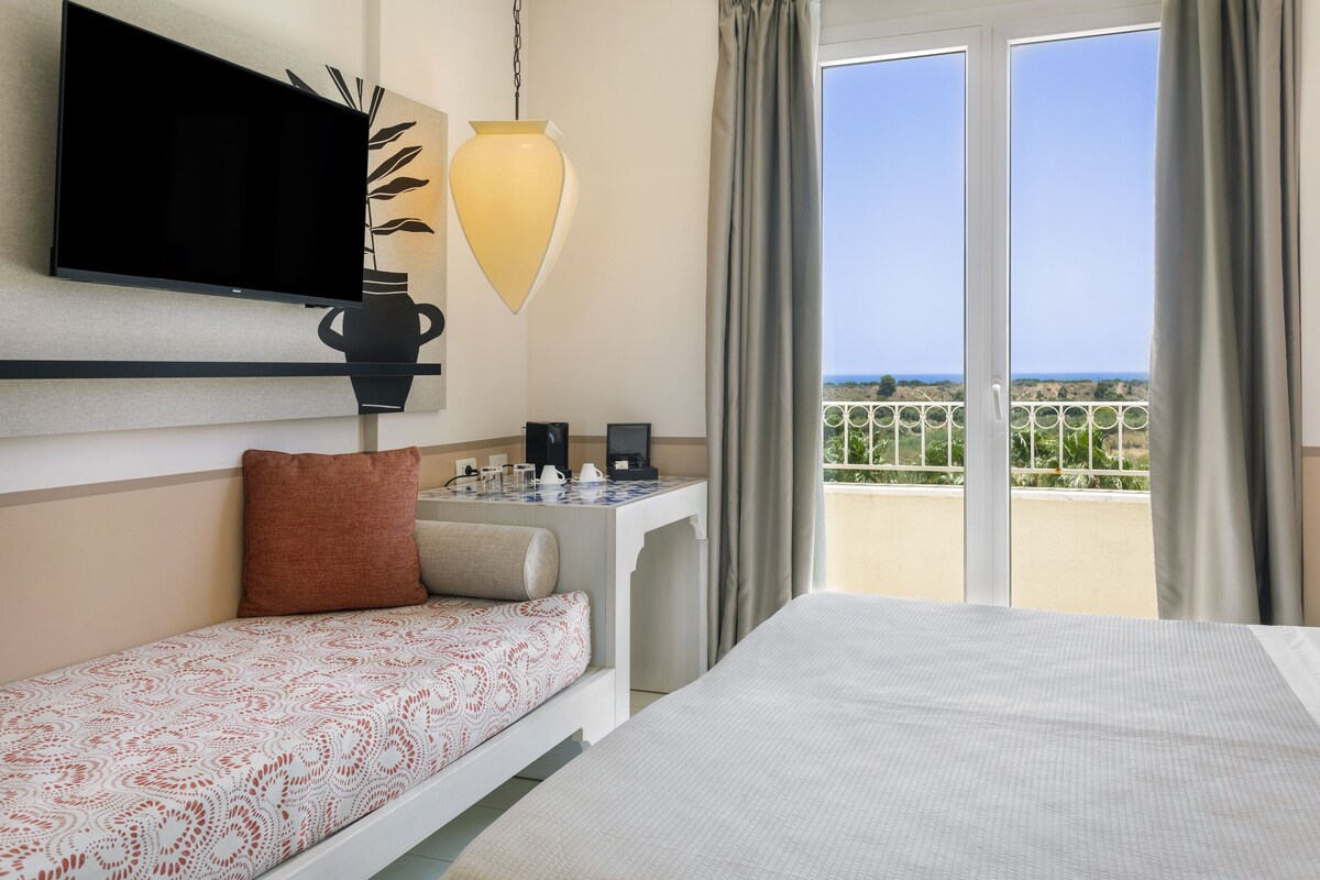 A hotel room with a bed, a sofa, a TV, a desk with a lamp, and a view of the sea through large windows.