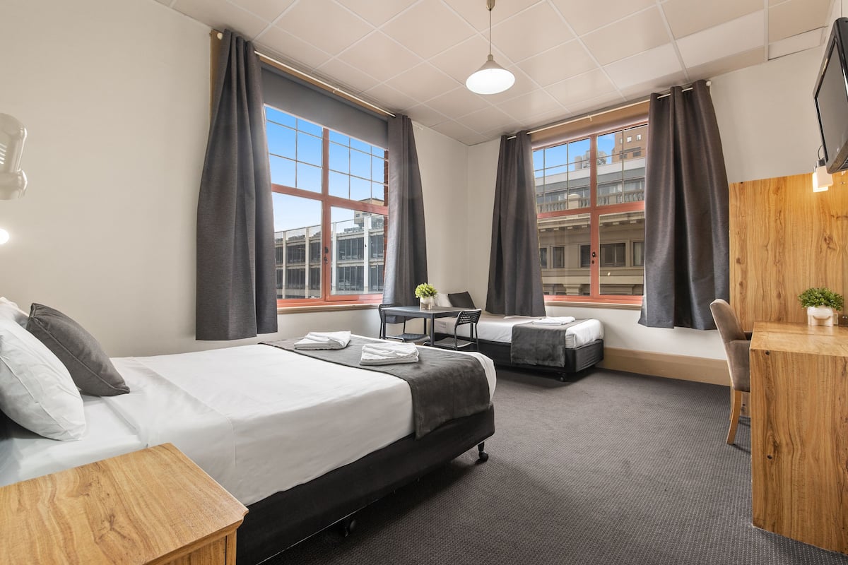 Hotellihuoneessa on suuri sänky, työpöytä, tuoli ja pieni pöytä. Huoneessa on kaksi ikkunaa, joissa on verhot, ja seinälle on asennettu televisio.