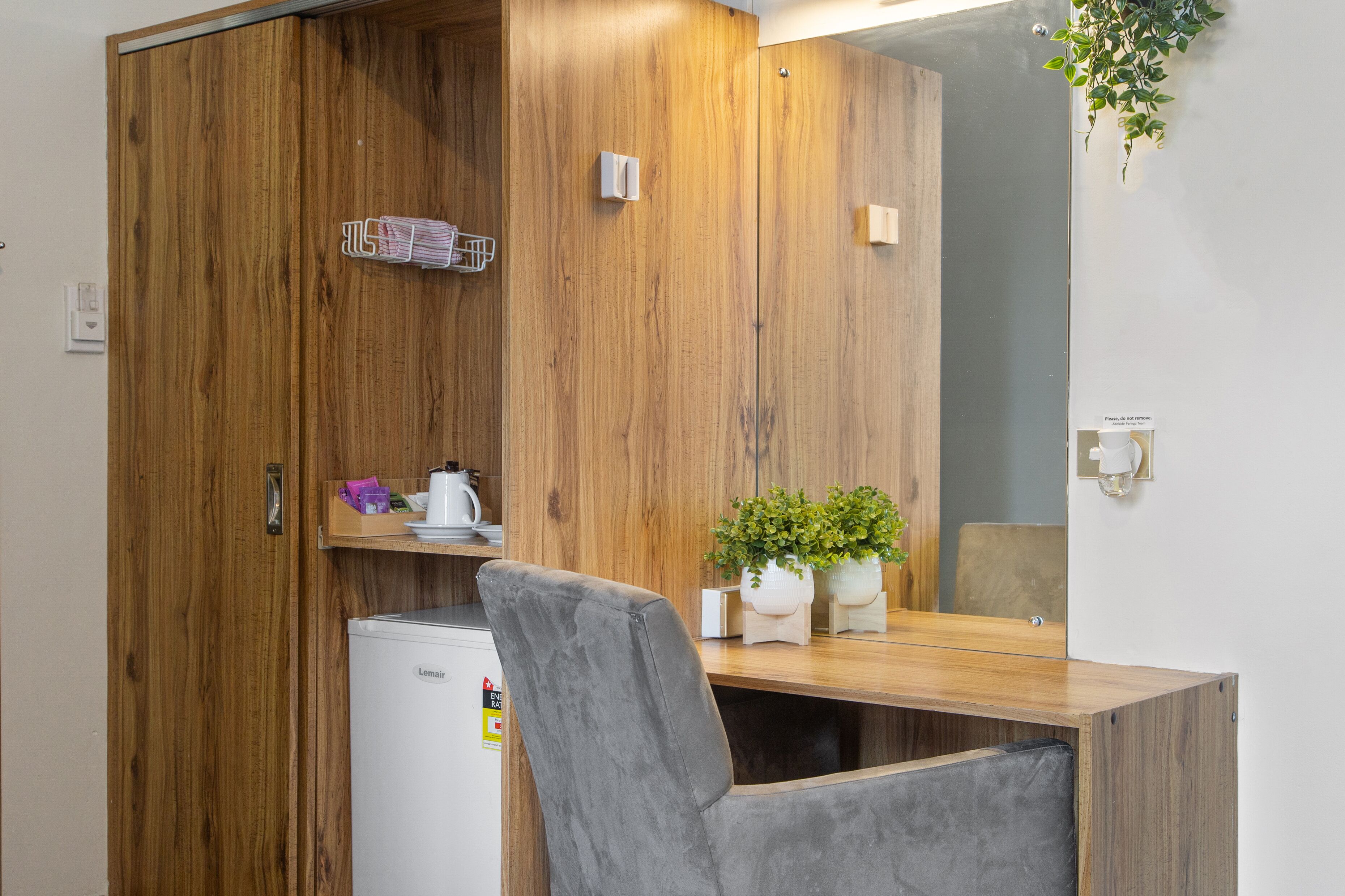 A compact room with a wooden wardrobe, a small refrigerator, a desk with a chair, and a potted plant.