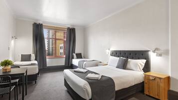 A hotel room with two beds, a desk, a chair, a small table with a plant, and a window with curtains.