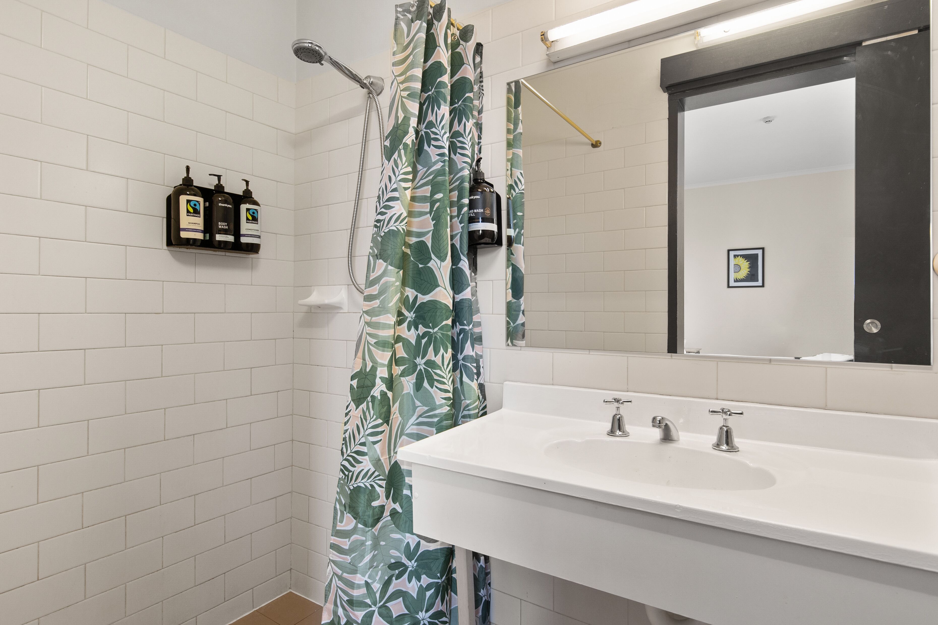A bathroom with white tiled walls, a white sink with two faucets, and a shower area with a green leaf-patterned shower curtain.