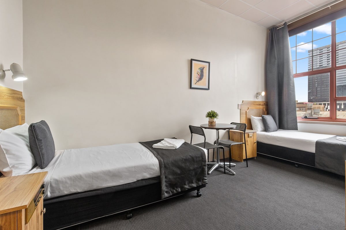Hotellihuone, jossa on kaksi erillistä sänkyä, pieni pöytä tuoleineen, työpöytä ja ikkuna, josta on näkymä kaupunkiin.