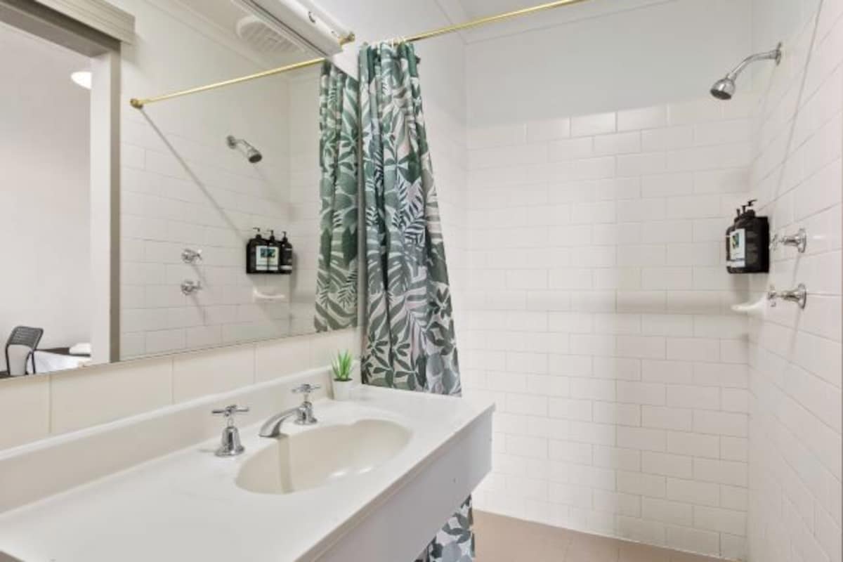 Un baño con un lavamanos blanco, un espejo, una ducha con cortina de estampado de hojas y una pared de azulejos blancos.