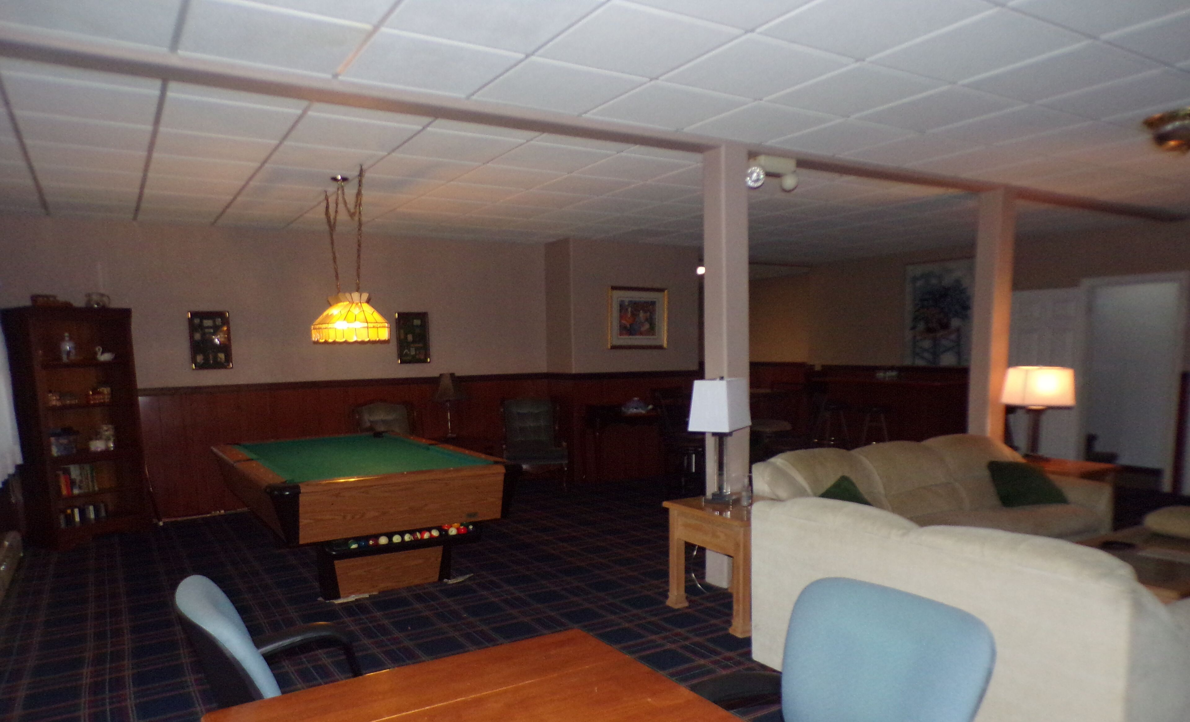 A billiards table, a pool cue, a sofa, a lamp, and a bookshelf in a room with a tiled floor.