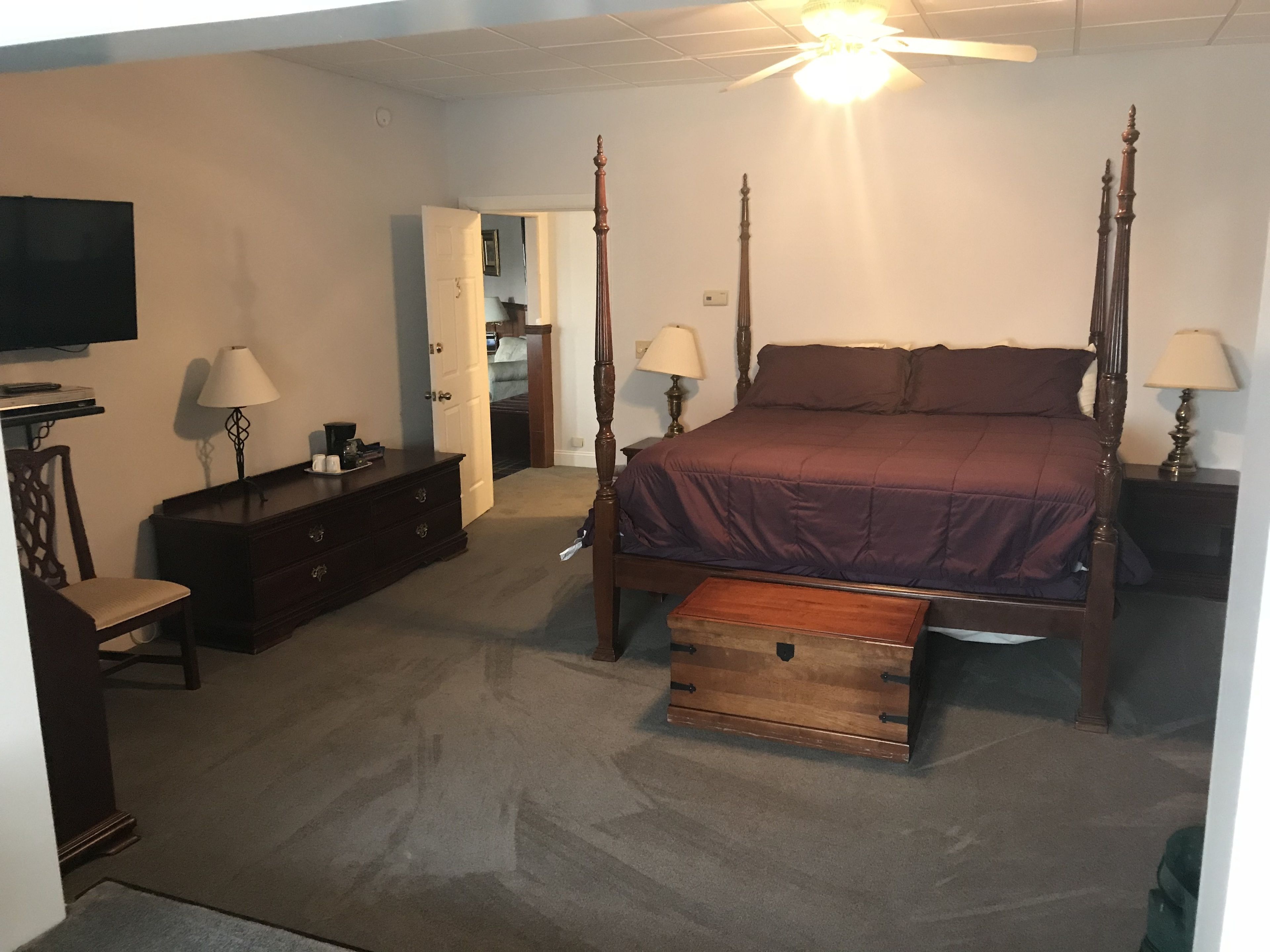 A bedroom with a four-poster bed, a dresser, a chair, a television on the wall, and a ceiling fan.