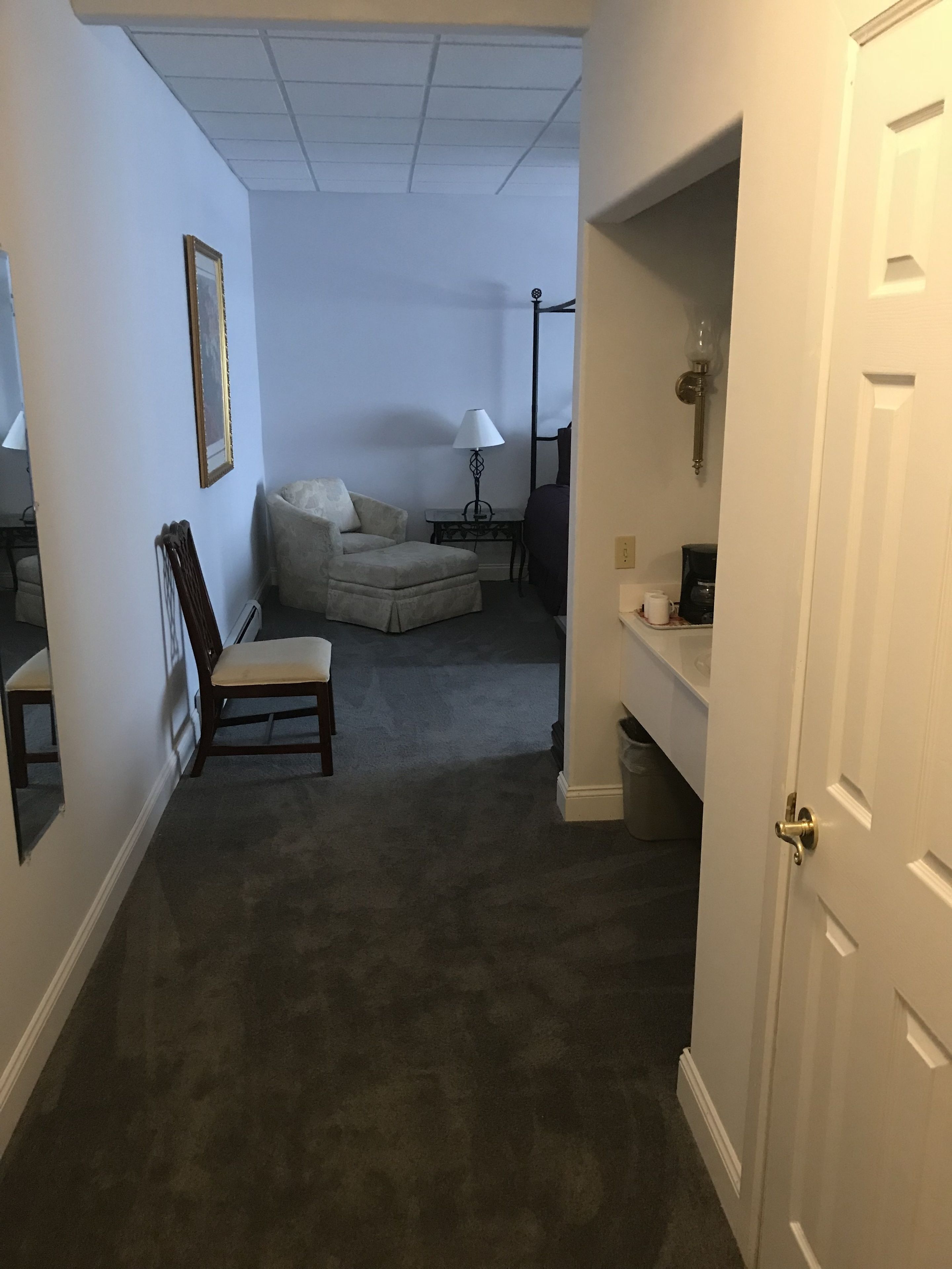 Um quarto com carpete escuro, uma cadeira, um sofá, um abajur e uma porta.