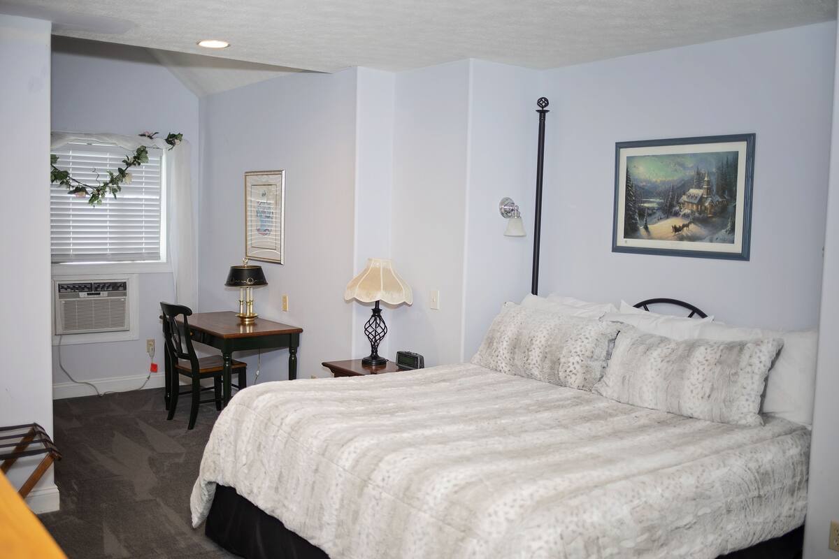 Une chambre à coucher avec un lit, un bureau, une chaise, une lampe, un tableau au mur et une fenêtre avec des stores.