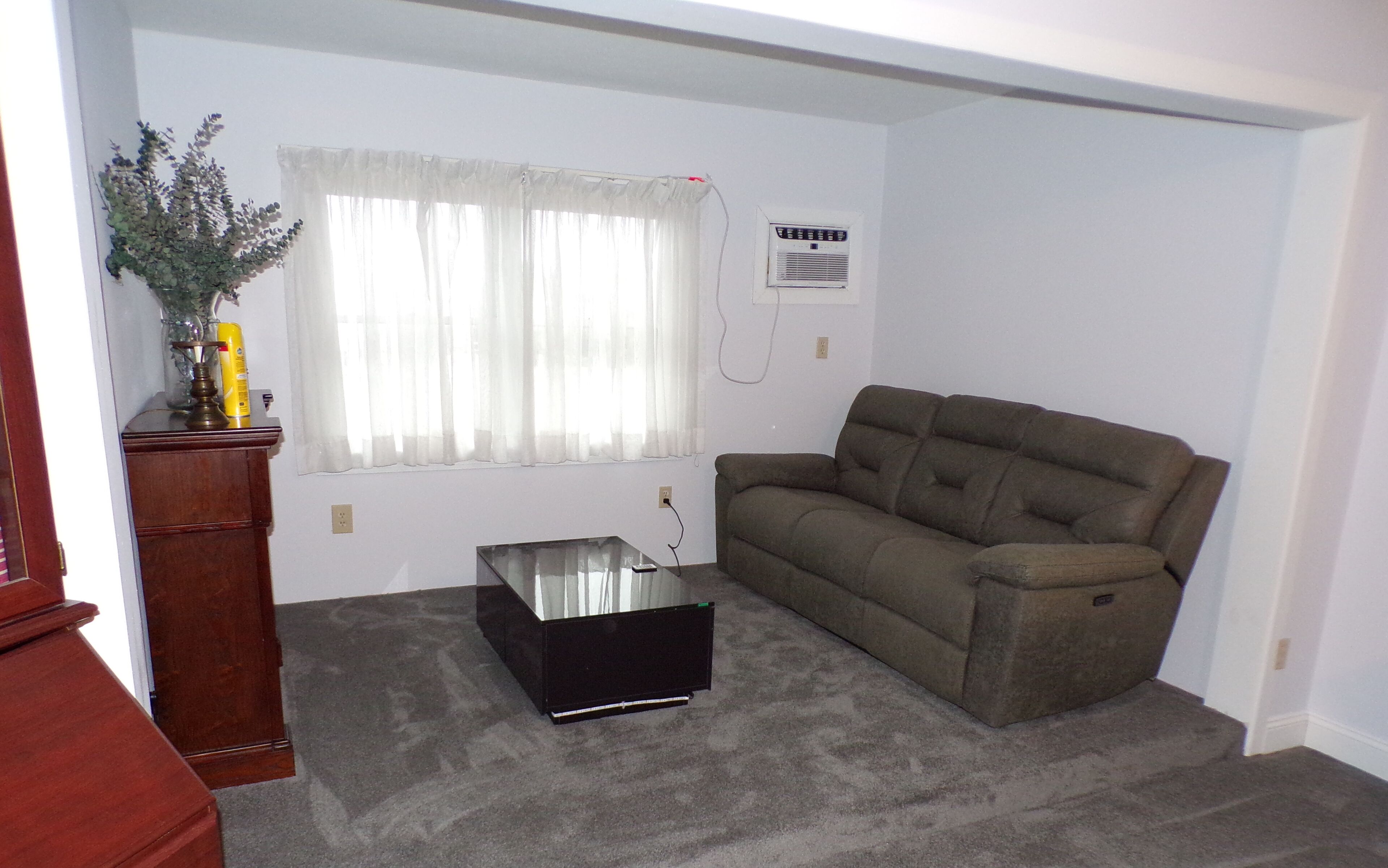 Una sala de estar con alfombra gris, un sofá marrón, un mueble de TV negro y una ventana con cortinas blancas.