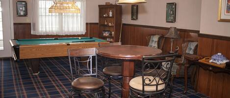 A pool table with four stools, a wooden table with chairs, a lamp, and a bookshelf.