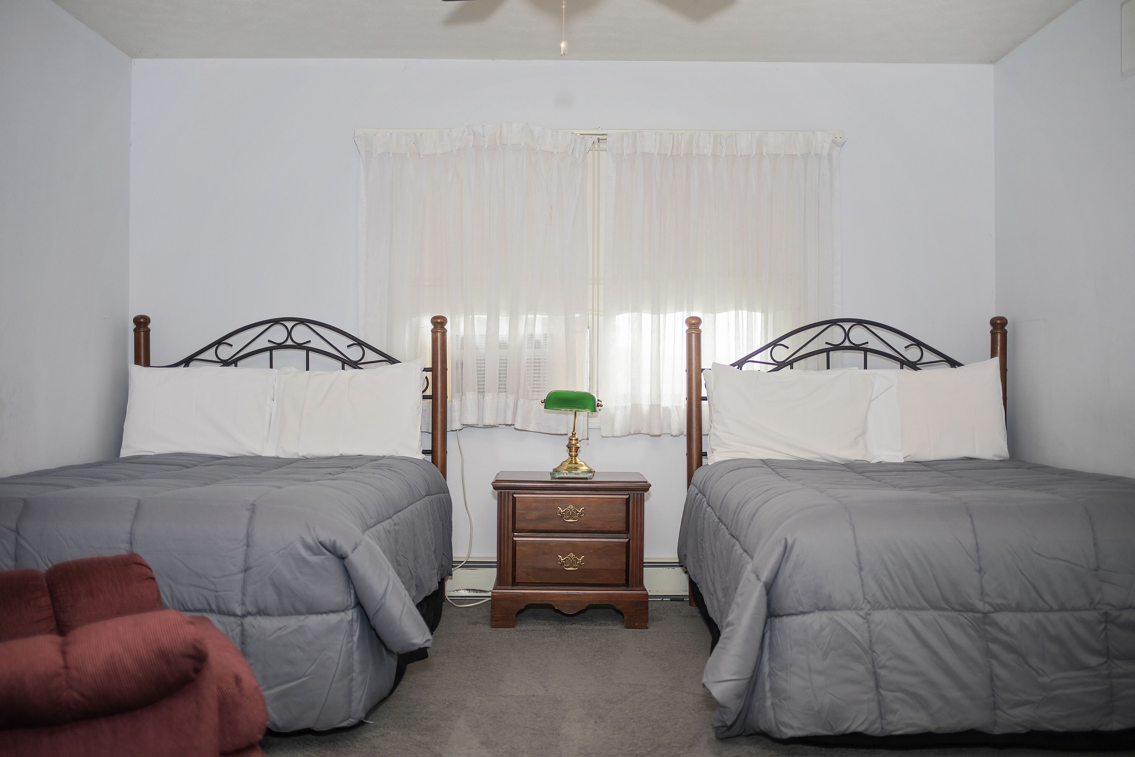A bedroom with two beds, a nightstand with a lamp, and a window with sheer curtains.