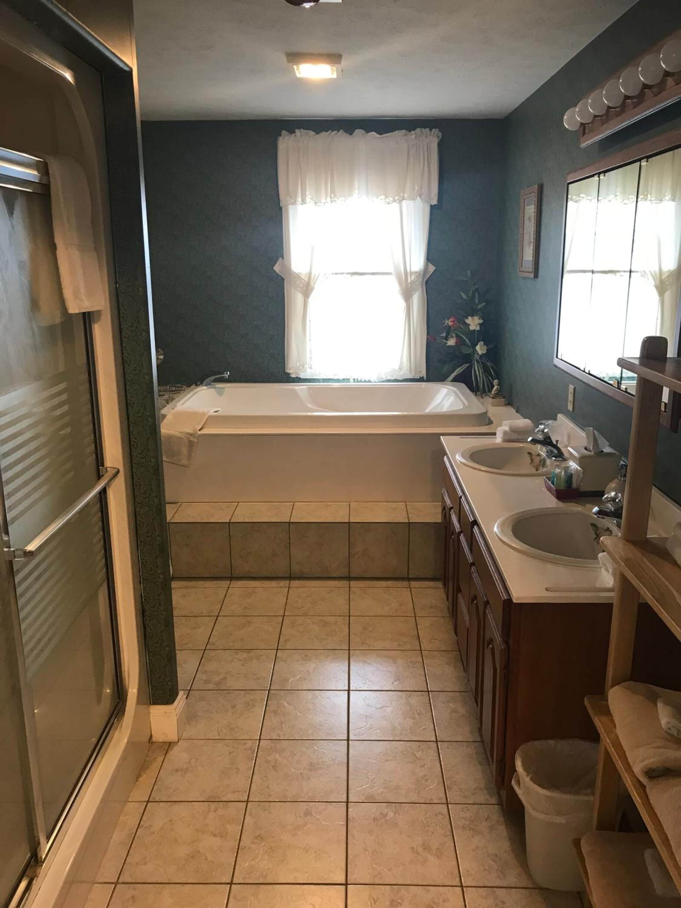A bathroom with a bathtub, two sinks, a mirror, and a window with curtains.