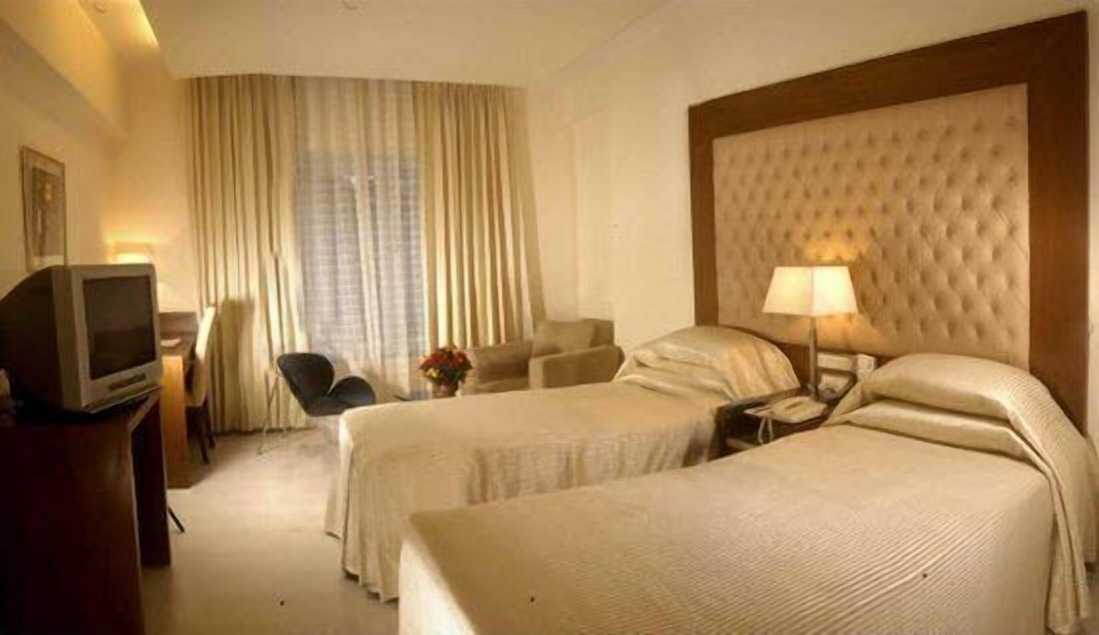 A hotel room with two beds, a TV, a desk with a lamp, and a large window with curtains.