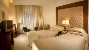 A hotel room with two beds, a TV, a desk with a lamp, and a large window with curtains. - The Chevron Hotel (Bengaluru)