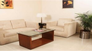 A living room with a beige sofa, a glass-top coffee table, a floor lamp, and a potted plant. - The Chevron Hotel (Bengaluru)