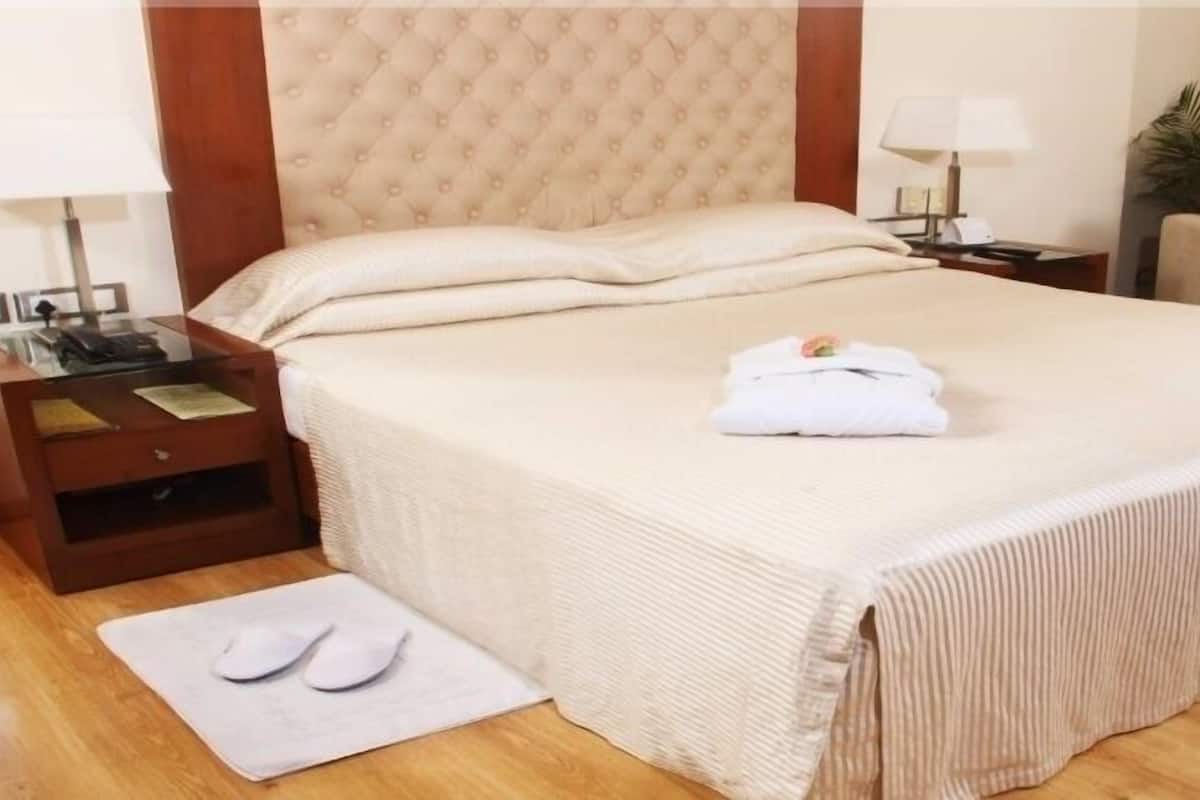 Una cama bien hecha con edredón blanco y almohadas, una mesita de noche con lámpara y teléfono, una silla y una pequeña mesa con un jarrón de flores.