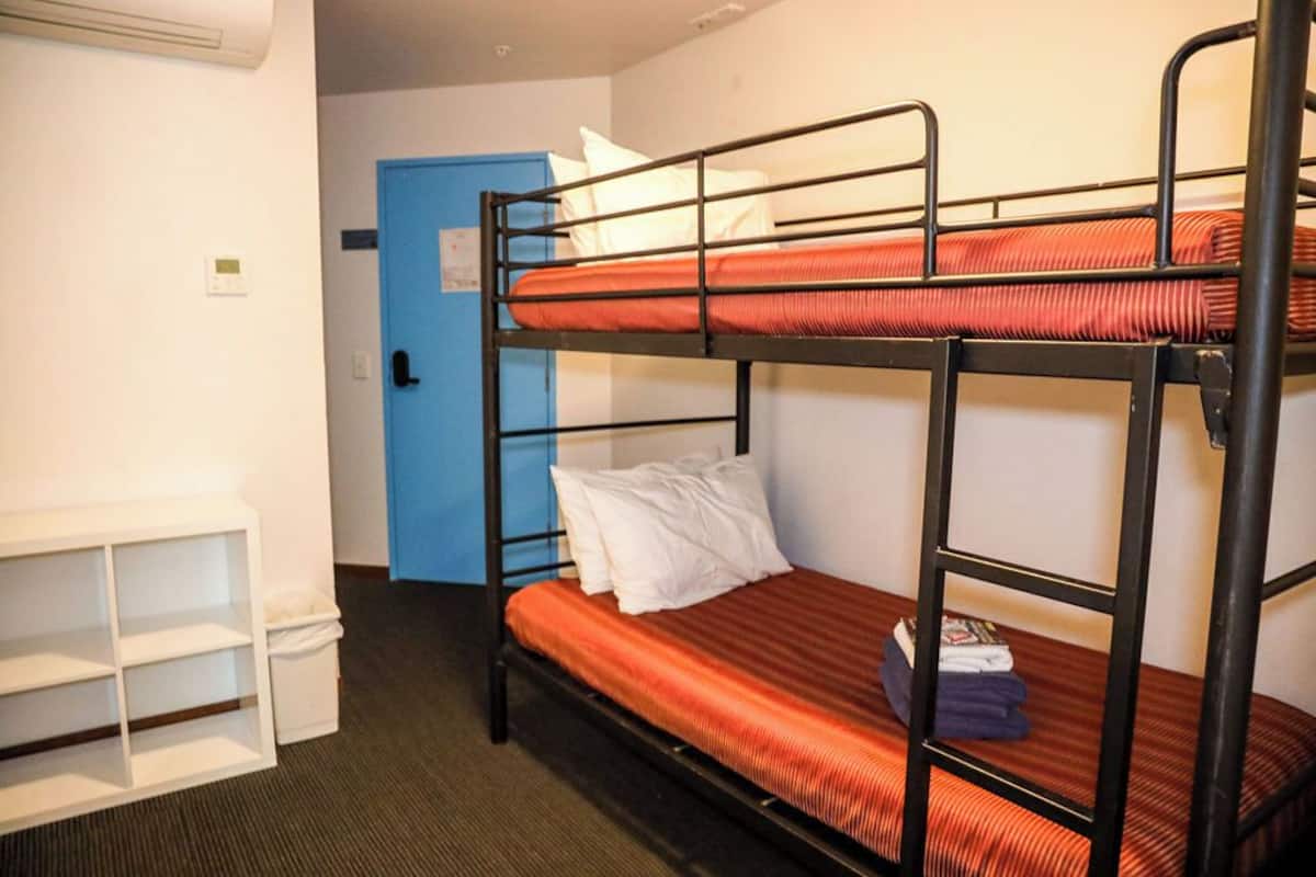 A bunk bed room with a blue door, a white shelf unit, and a small trash bin.
