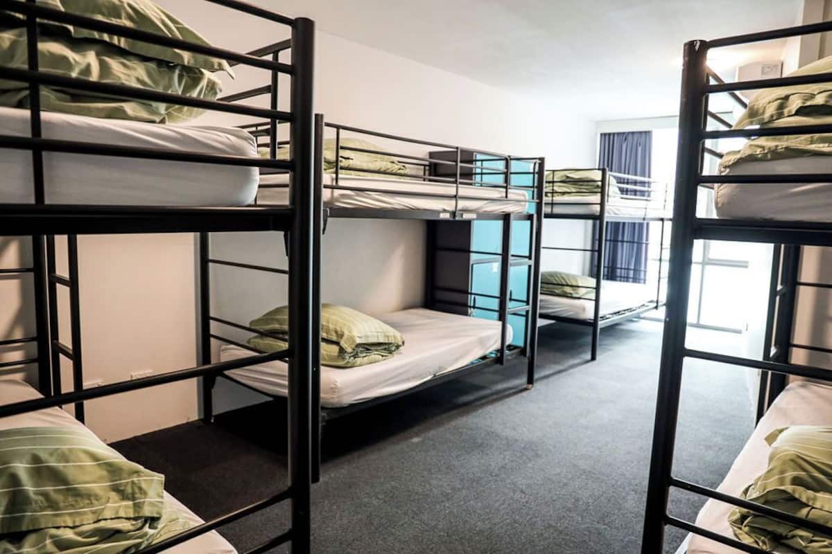 A dormitory room with bunk beds arranged in a row, a window with curtains, and a door in the background.
