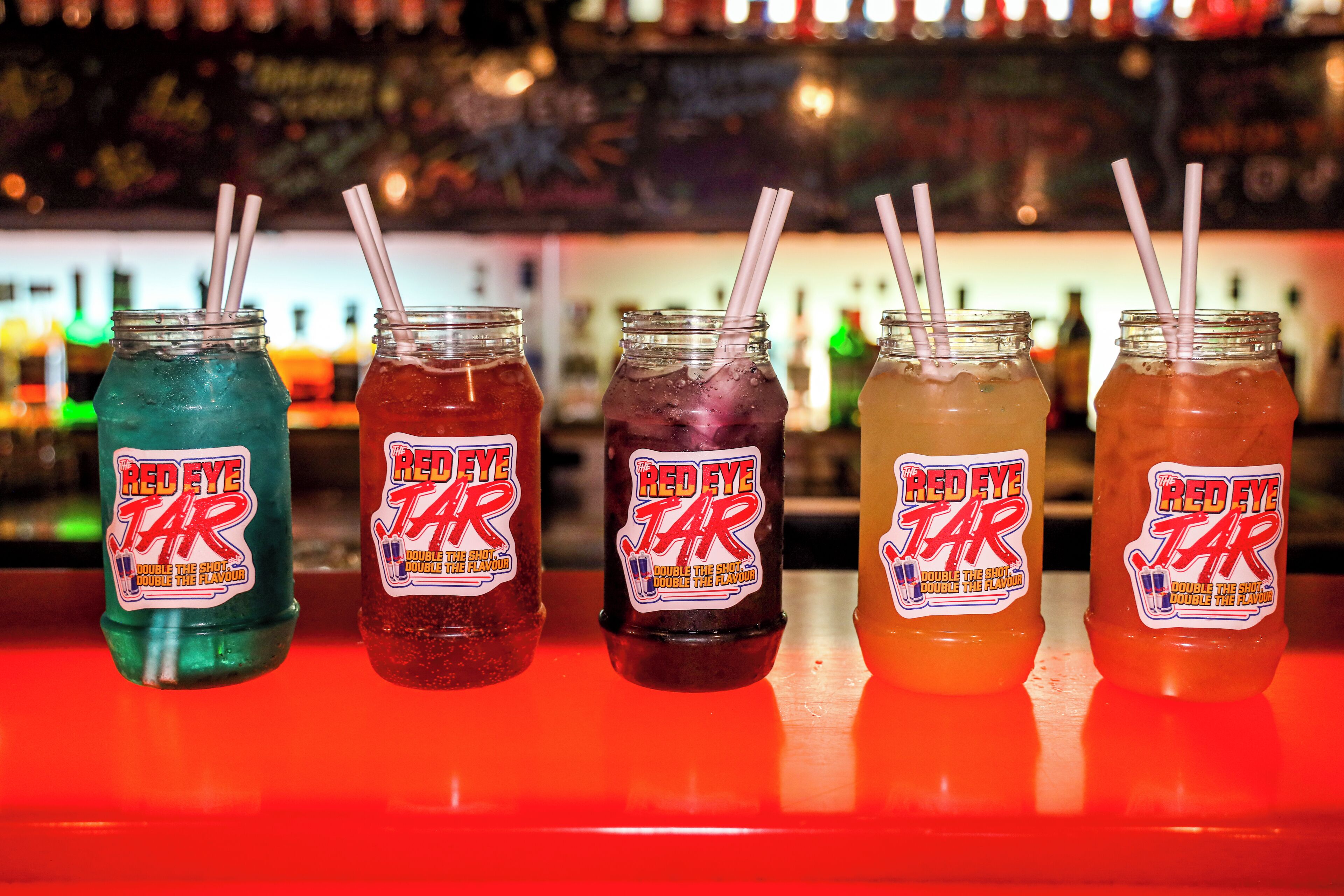 Cinco bebidas Red Eye Tar de varios colores con pajitas, exhibidas sobre una barra.
