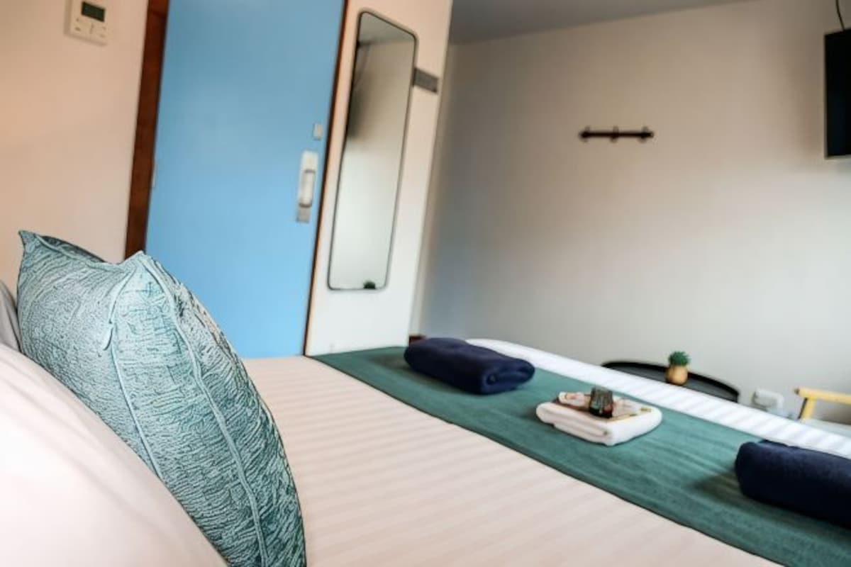 A hotel room with a bed, a green bedspread, a small table with a towel and a plant, a mirror on the wall, and a door leading to another room.
