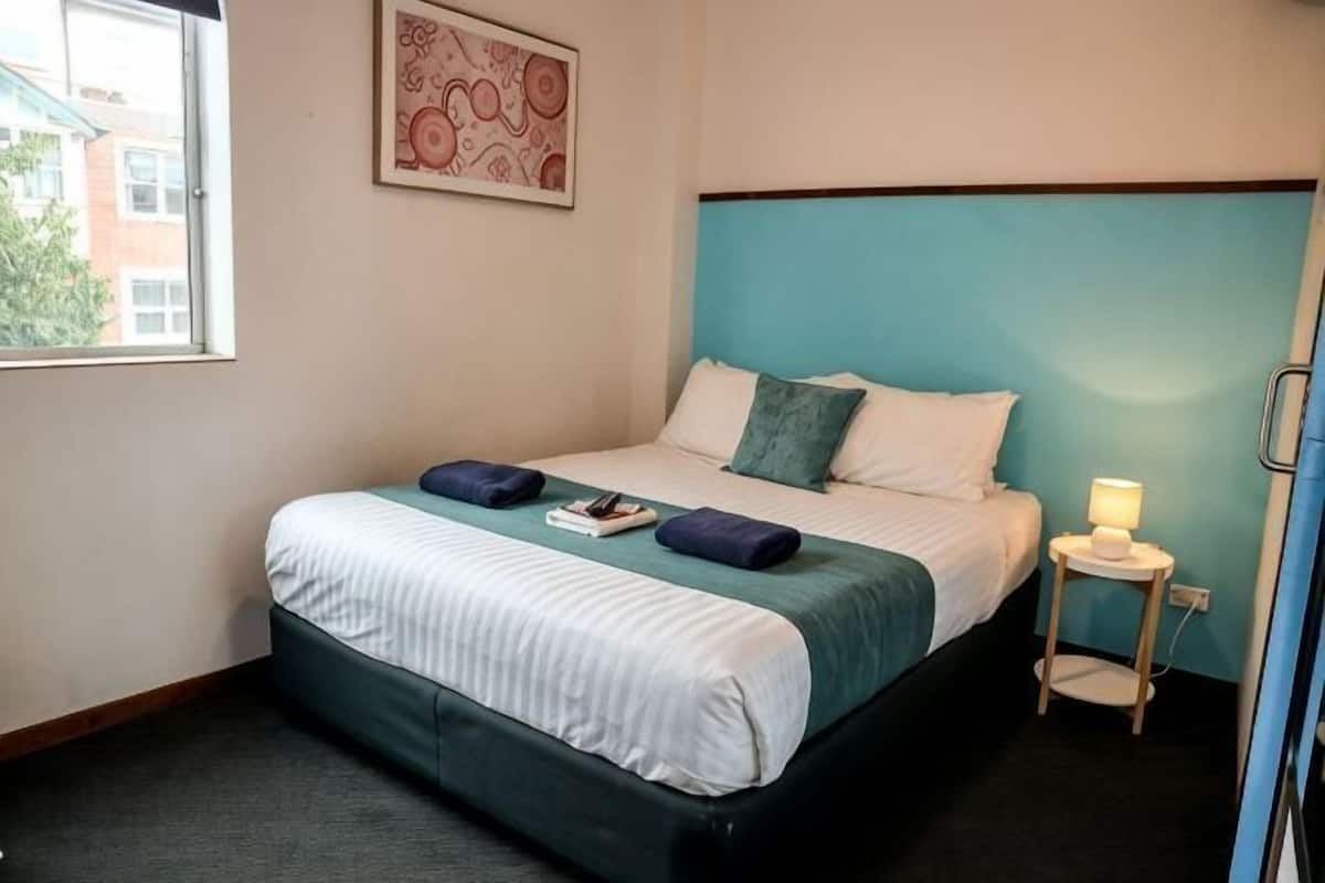 A hotel room with a bed, a bedside table with a lamp, a window, and a wall-mounted artwork.