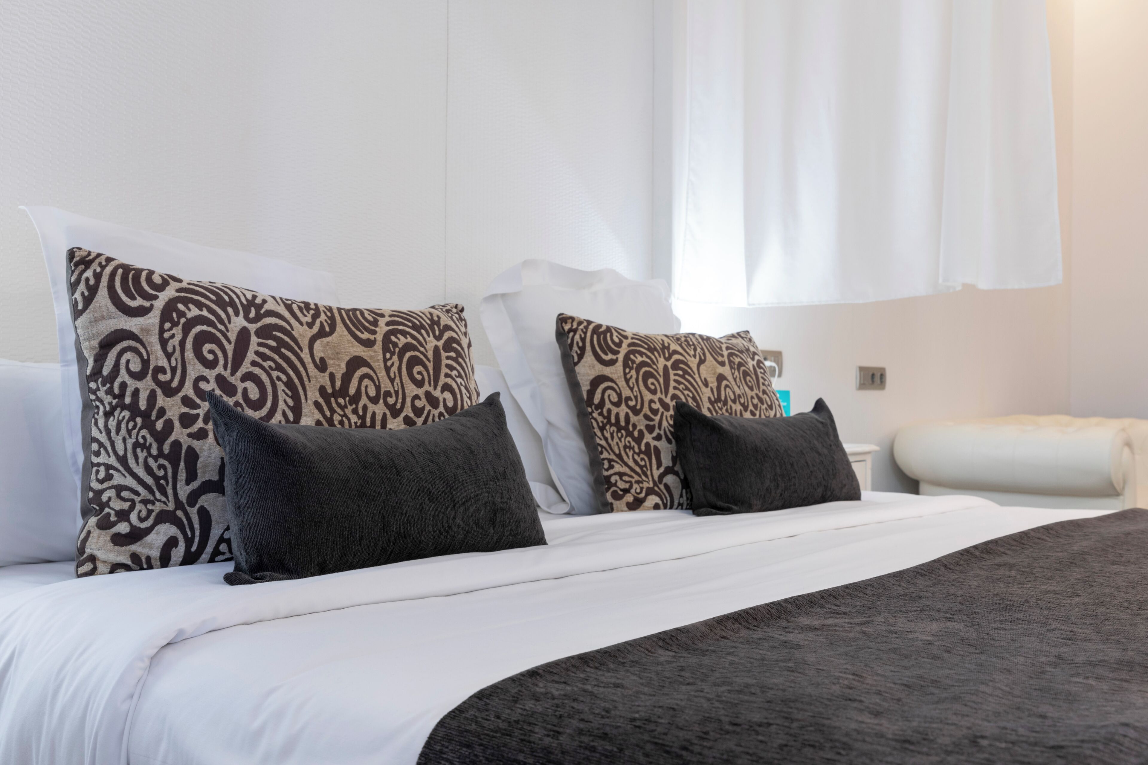 Ein ordentlich bezogenes Bett mit gemusterten Kissen, einem weißen Kopfteil und einer weißen Wand.