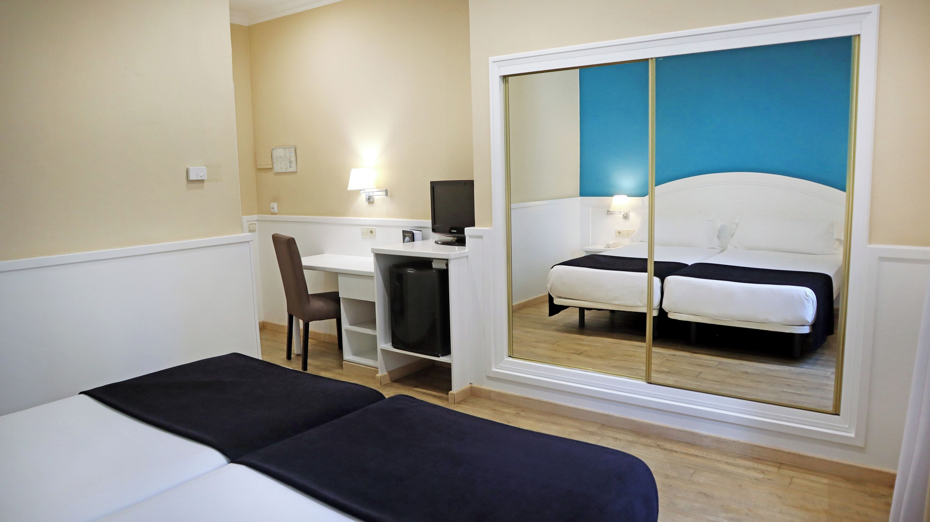 Une chambre d’hôtel comprenant un lit, un bureau avec un ordinateur, une chaise et une armoire avec un miroir.
