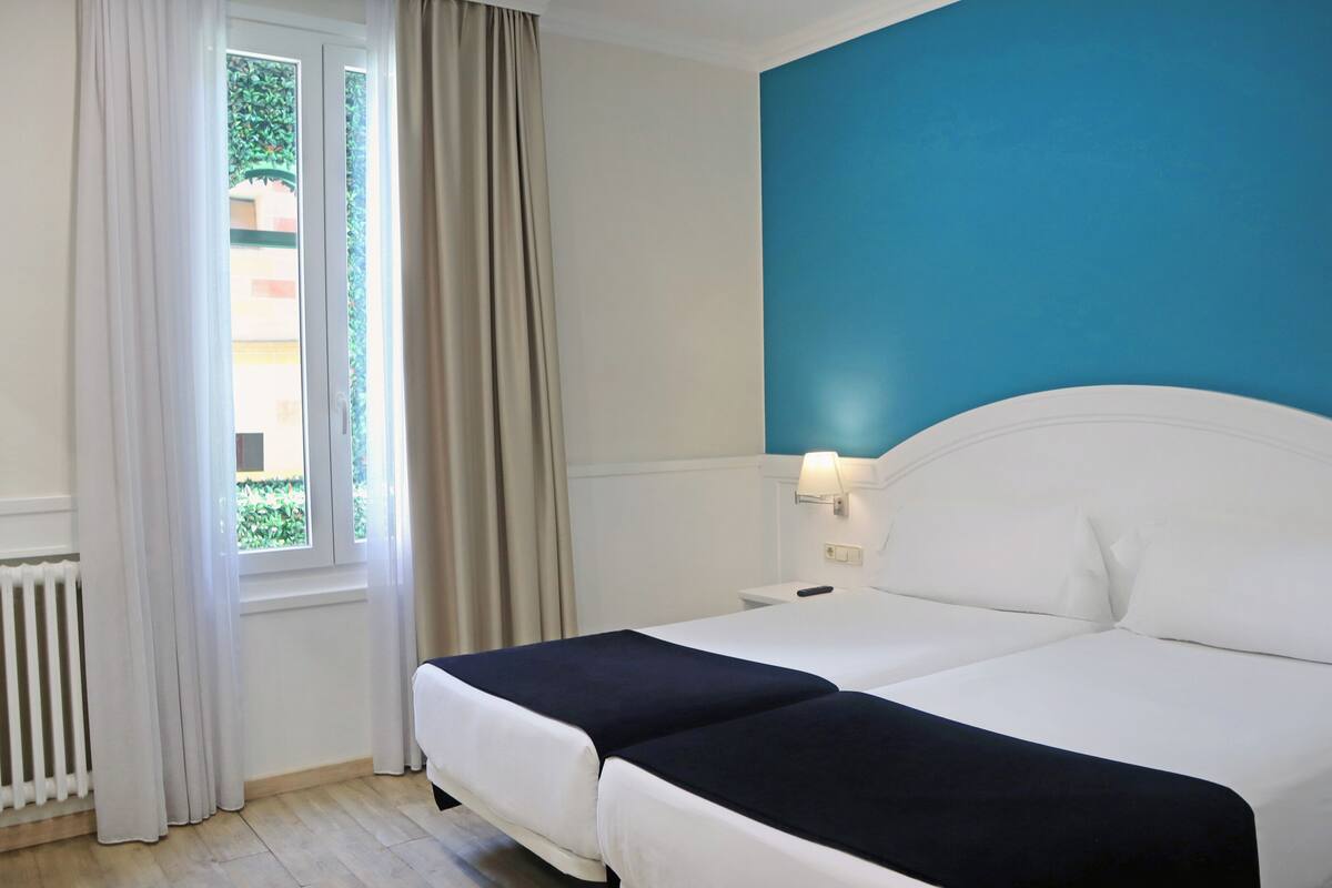 Quarto de hotel com cama, mesa de cabeceira, abajur, janela com cortinas e radiador.