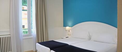 A hotel room with a bed, a nightstand, a lamp, a window with curtains, and a radiator.